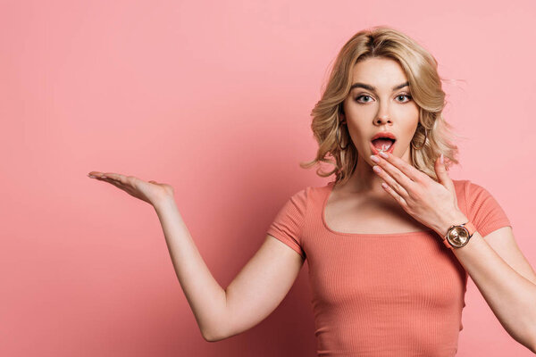 shocked girl covering mouth with hand while pointing with finger on pink background