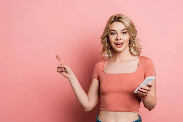 Smiling Girl Pointing Finger While Holding Smartphone Pink Background — ストック写真