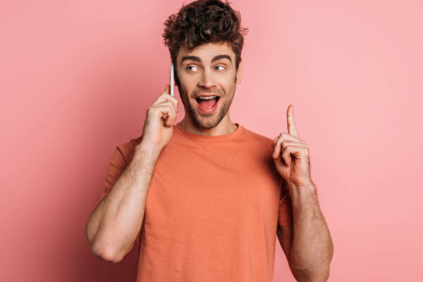 excited young man showing idea gesture while talking on smartphone on pink background