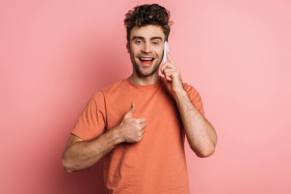 happy young man showing thumb up while talking on smartphone on pink background
