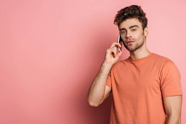 serious young man looking at camera while talking on smartphone on pink background
