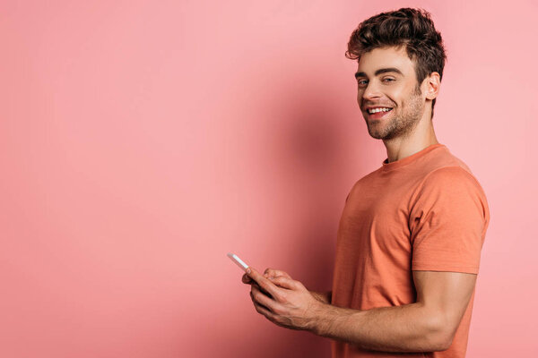 cheerful young man smiling at camera while chatting on smartphone on pink background