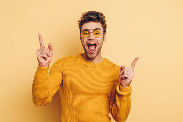 excited young man pointing with fingers while looking at camera on yellow background
