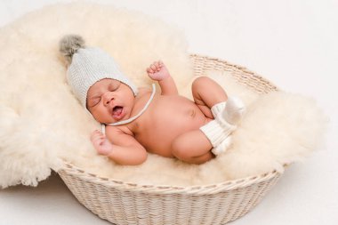  newborn mixed race baby in knitted hat yawning in basket on white clipart