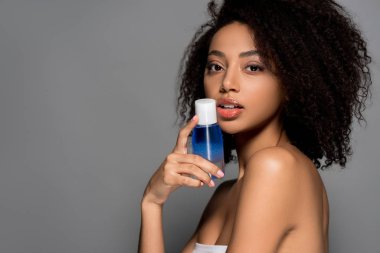Seksi Afrikalı Amerikalı kız elinde bir şişe makyaj temizleyicisi var, gri renkte izole edilmiş.
