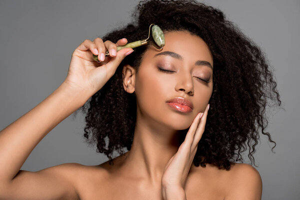 beautiful african american girl with closed eyes using jade roller, isolated on grey