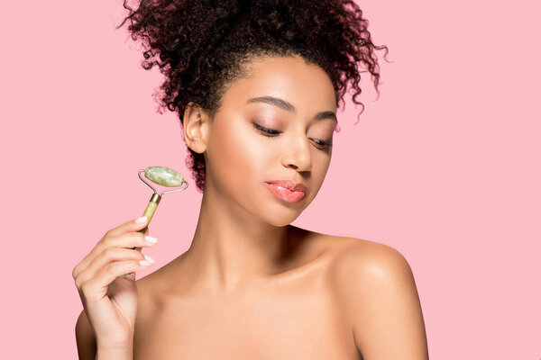 happy naked african american young woman using jade roller, isolated on pink
