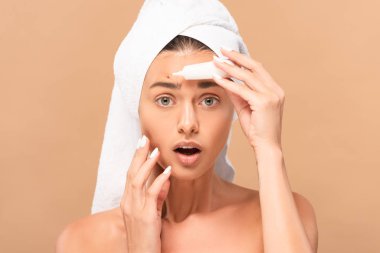 shocked and naked woman in towel holding treatment cream near pimple on face isolated on beige  clipart
