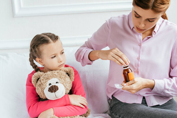 mom giving syrup to sick daughter with teddy bear