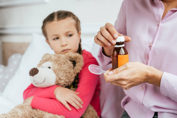 cropped view of mother giving syrup to ill daughter with teddy bear