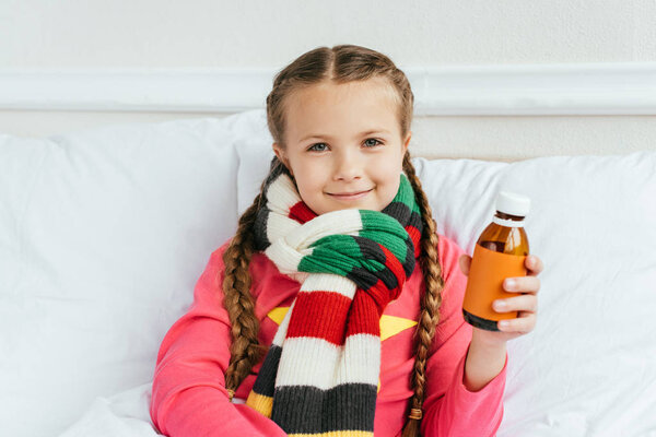 smiling ill kid in scarf holding syrup and sitting on bed