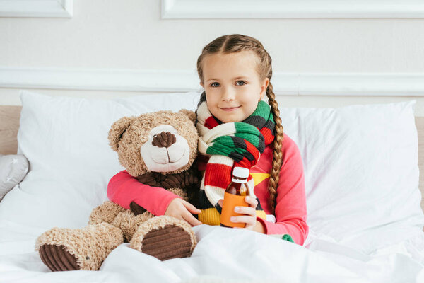 smiling sick child in scarf holding syrup and sitting on bed with teddy bear
