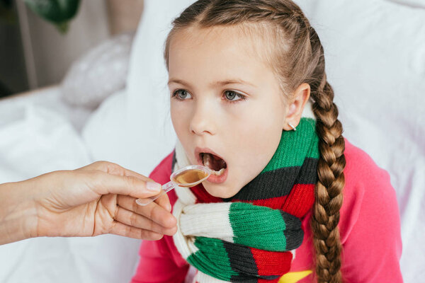 mom giving syrup to ill daughter with scarf in bed