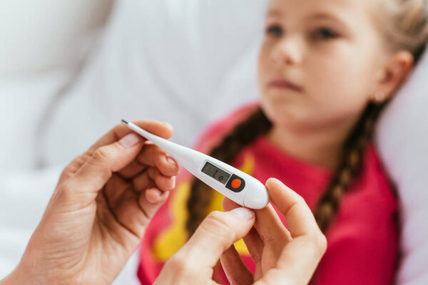 selective focus of mother looking at thermometer with sick daughter in bed