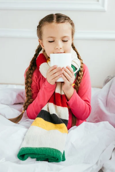ベッドの上で熱い飲み物を持っているスカーフの病気の子供 — ストック写真