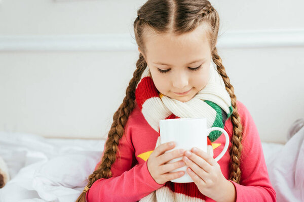 cute ill kid in scarf holding cup of hot drink while sitting on bed