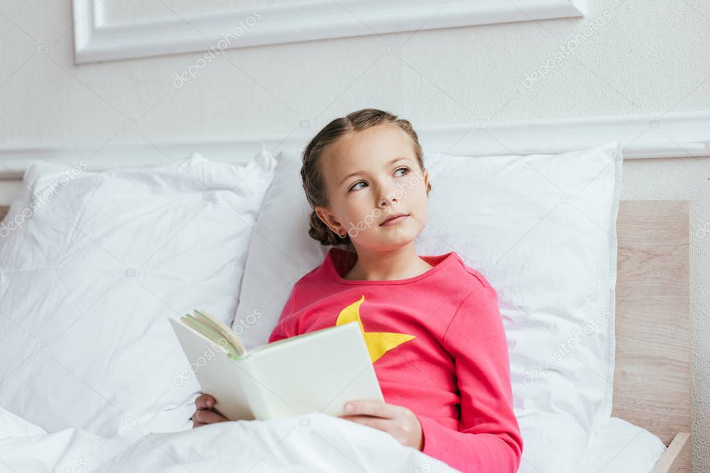 pensive kid reading book while sitting on bed