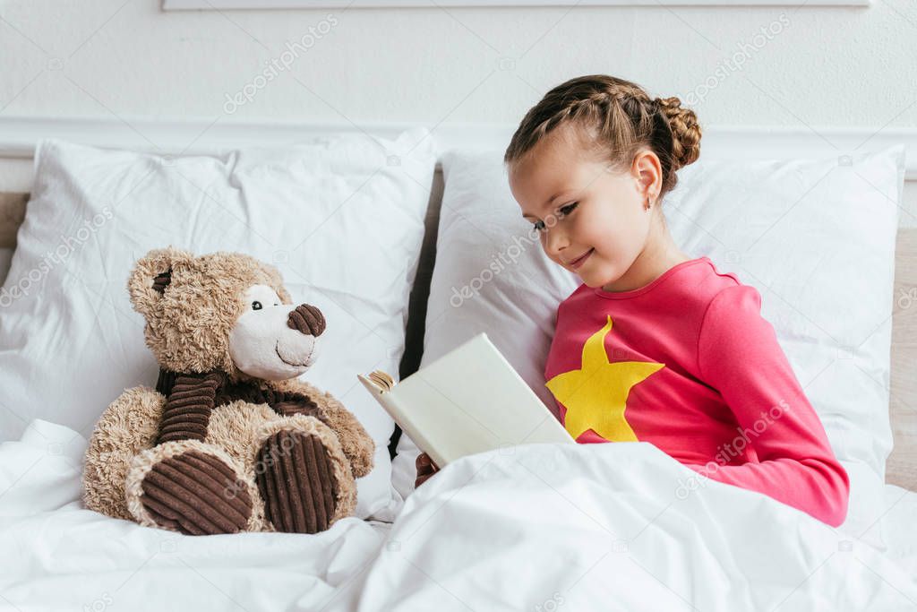 adorable child reading book to teddy bear while sitting on bed
