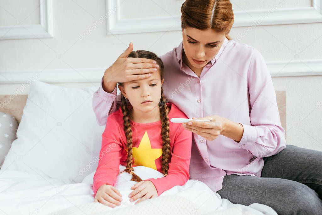 mother looking at thermometer while touching forehead of ill daughter with fever