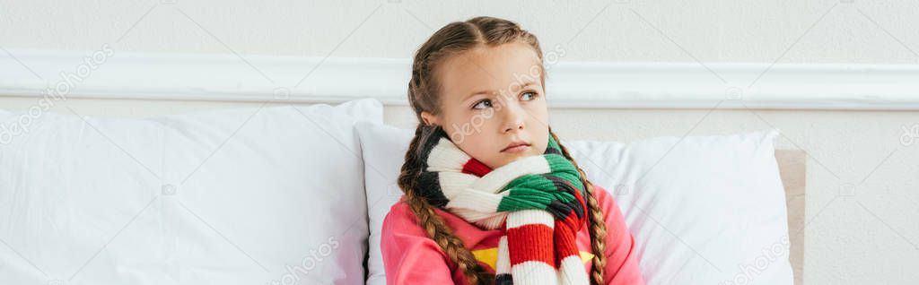 panoramic shot of upset sick kid in scarf sitting on bed