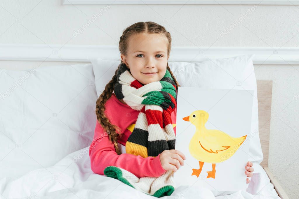 smiling sick kid in scarf holding painting with duck on bed
