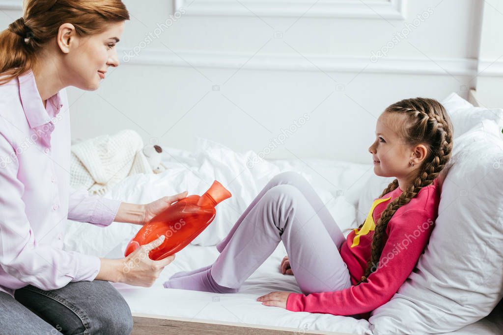smiling mother giving hot water bottle to ill daughter sitting on bed