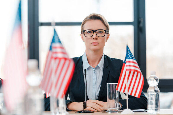selective focus of attractive diplomat in eyeglasses near american flags on table 