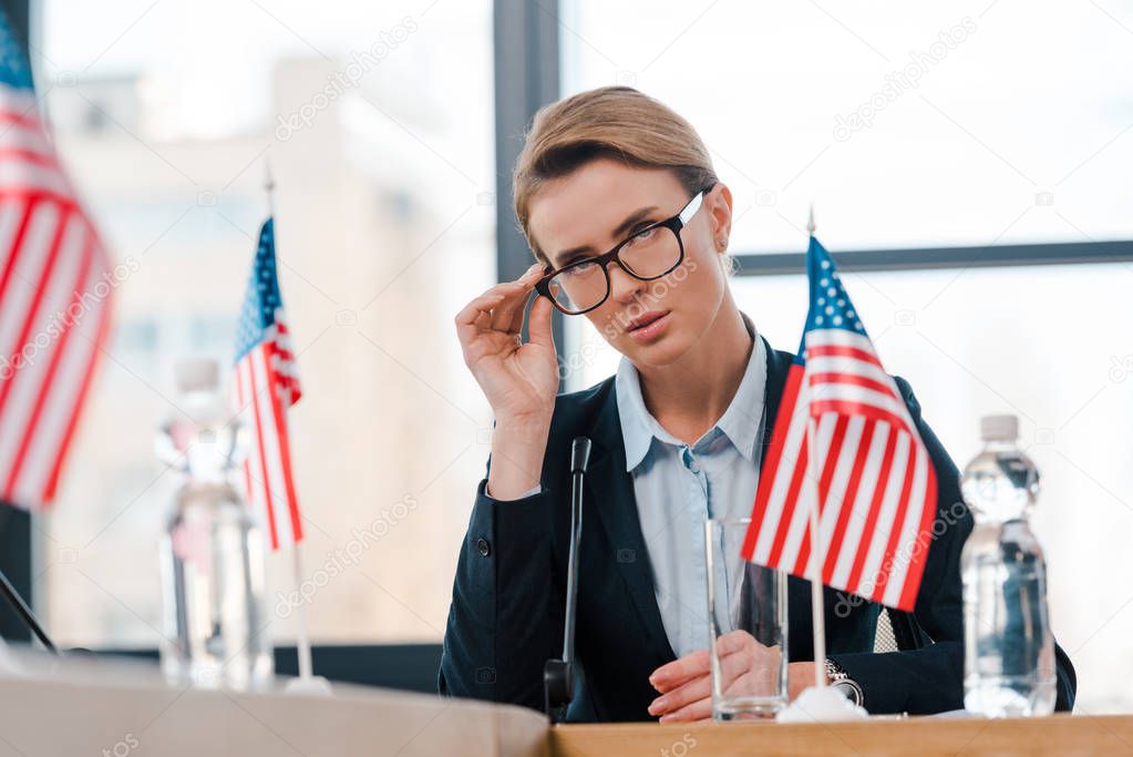 selective focus of beautiful diplomat touching eyeglasses near american flags