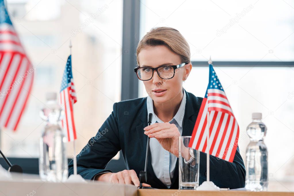 selective focus of beautiful diplomat in eyeglasses touching microphone near american flags