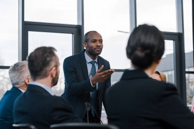 selective focus of african american diplomat gesturing near businessmen and businesswomen  clipart
