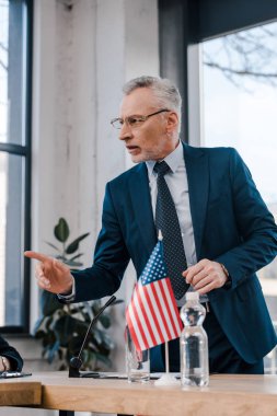 Gözlüklü, Amerikan bayrağının yanında konuşurken parmağıyla işaret eden sakallı diplomatın seçici odağı. 