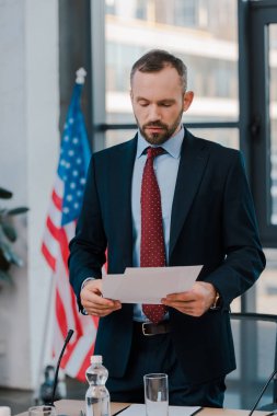 Takım elbiseli sakallı diplomatın seçici odak noktası Amerikan bayrağının yanında kağıtları tutuyor. 