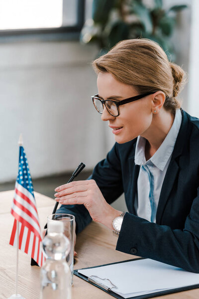 selective focus of attractive diplomat in eyeglasses touching microphone near clipboard and american flag 