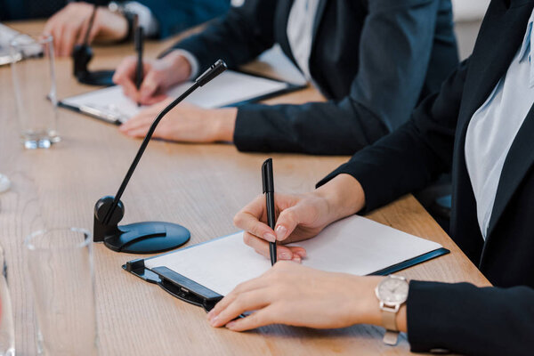 cropped view of businesswomen writing near microphones and glasses on table 