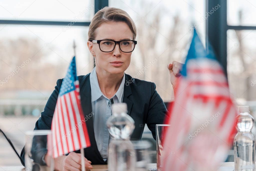 selective focus of patriotic woman in eyeglasses with clenched fist near american flags 