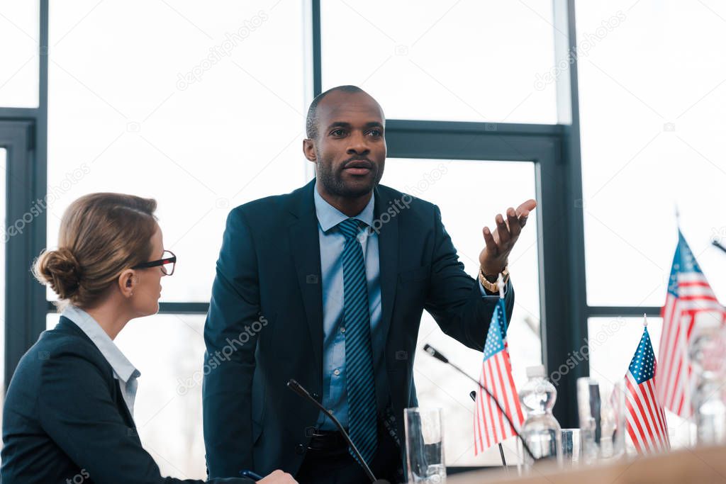 selective focus of attractive diplomat looking at african american man gesturing while talking near flags of america 
