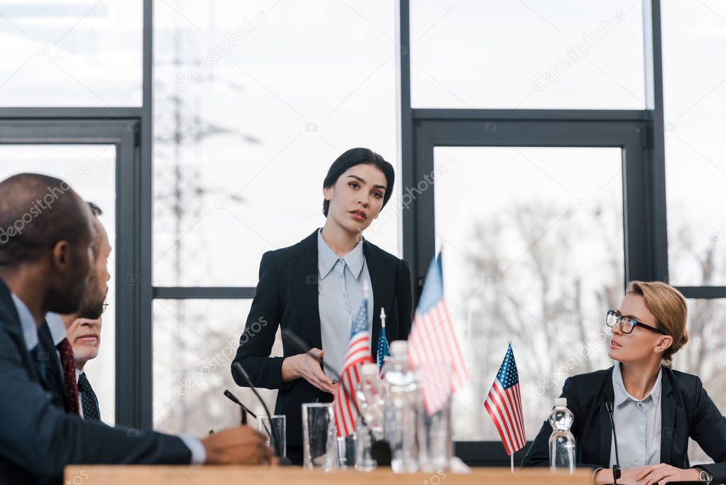 selective focus of attractive speaker talking near multicultural diplomats and american flags 