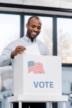  Neşeli Afrikalı Amerikalı adam oy veriyor ve oy pusulasını Amerika bayrağıyla kutuya koyuyor.