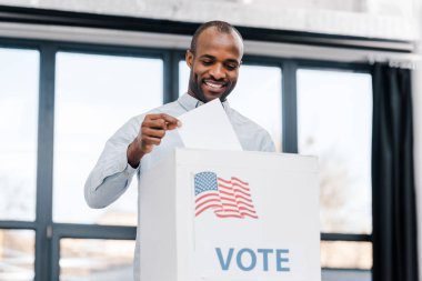  Mutlu Afrikalı Amerikalı adam oy veriyor ve oy pusulasını Amerika bayrağıyla kutuya koyuyor.