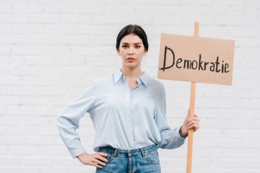 Elinde demokrat yazılı bir pankart tutan ve elleri tuğla duvarın yanında duran bir kadın. 
