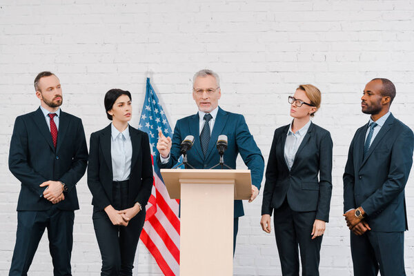 bearded speaker in glasses pointing with finger near microphones and multicultural diplomats 