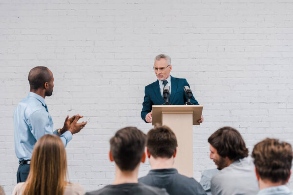 happy speaker looking at african american businessman talking and gesturing near journalists 