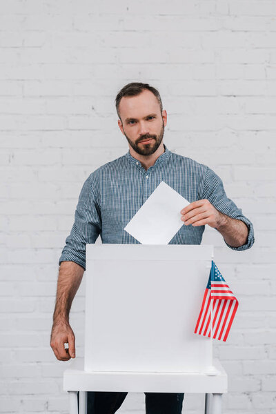bearded and handsome voter putting blank ballot in voting box near american flag 