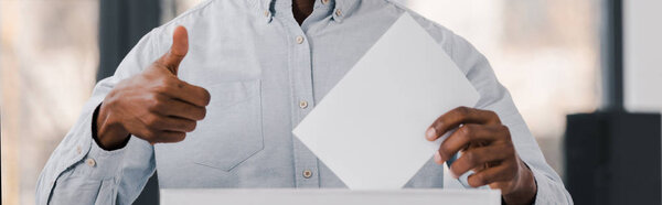 панорамный снимок африканского избирателя, держащего пустой бюллетень и показывающего большой палец вверх
 