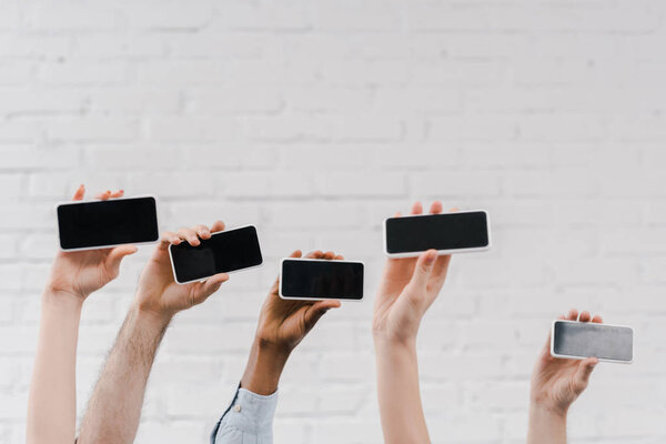 cropped view of multicultural people holding smartphones with blank screen near brick wall 
