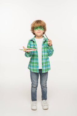 full length view of cute boy with gecko mask painted on face holding palette and paintbrush on white background clipart