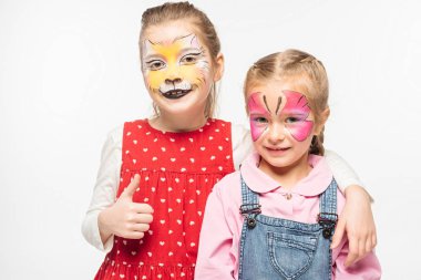 adorable kid with cat muzzle painting on face showing thumb up while embracing friend with painted butterfly mask isolated on white clipart