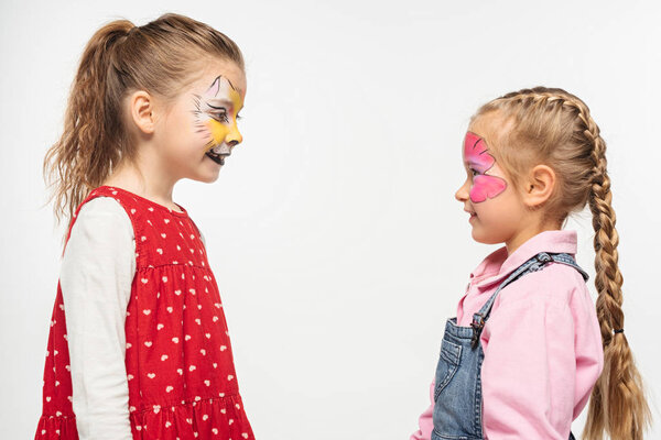 side view of smiling friends with cat muzzle and butterfly paintings on faces looking at each other isolated on white