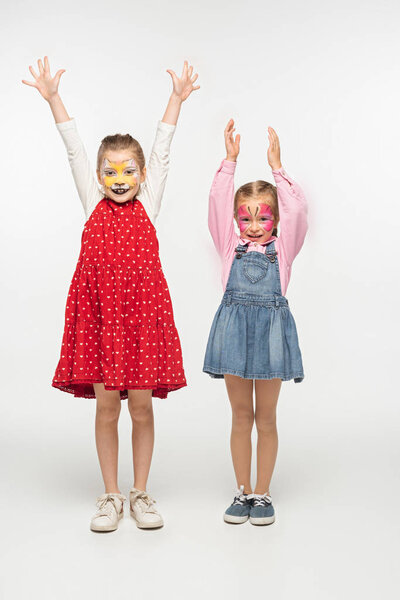 full length view of adorable children with cat muzzle and butterfly paintings on faces standing with raised hands on white background