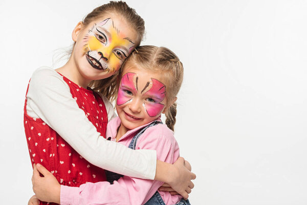 cute children with cat muzzle and butterfly paintings on faces embracing while looking at camera isolated on white